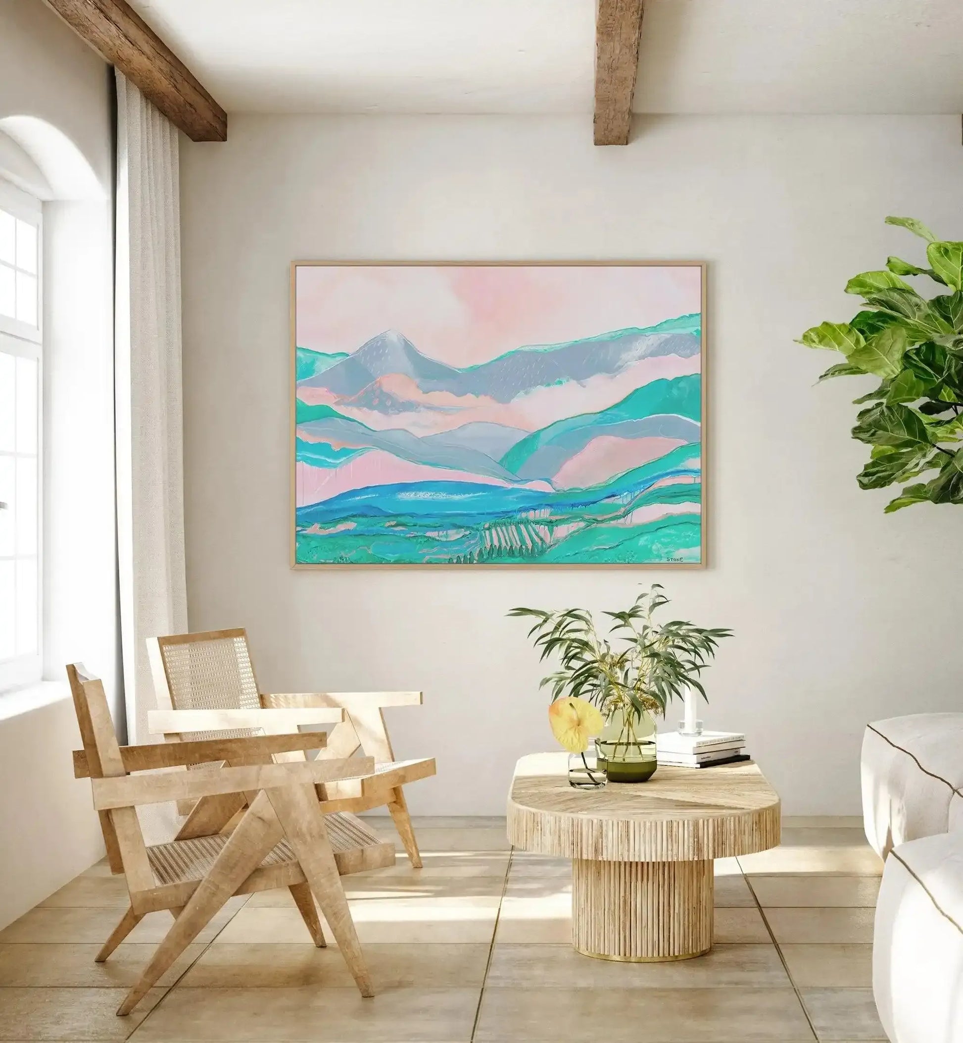 Lifestyle shot of a framed canvas art print displayed in a bright, minimalist living room with natural wood accents. The print features an abstract landscape with rolling mountains and a valley, rendered in soft pastel shades of dusty pink, sky blue, and mint green, with a natural oak frame. The room includes two light wood chairs with woven cane backs, a round wooden coffee table with a fluted base, and a large green plant in the corner.