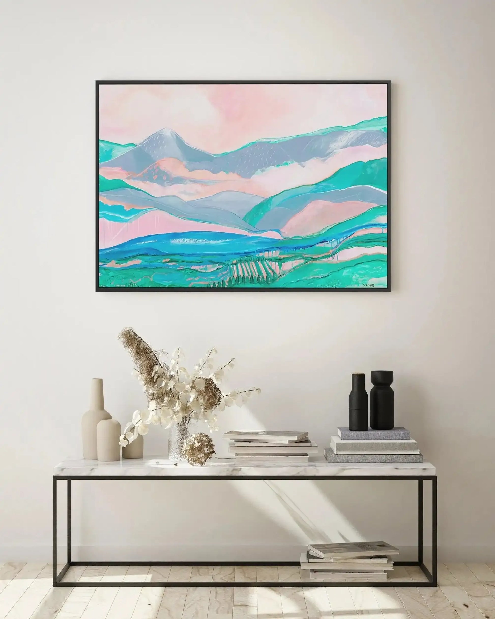 Lifestyle shot of a framed canvas art print displayed above a marble-topped console table in a minimalist room. The abstract landscape print features rolling hills and mountains in shades of soft pink, pale blue, and teal green, with a bright blue river or lake in the foreground and a hint of a vineyard. The print is in a thin black frame. The console table is styled with cream-colored ceramic vases, dried floral arrangements, and stacks of books.