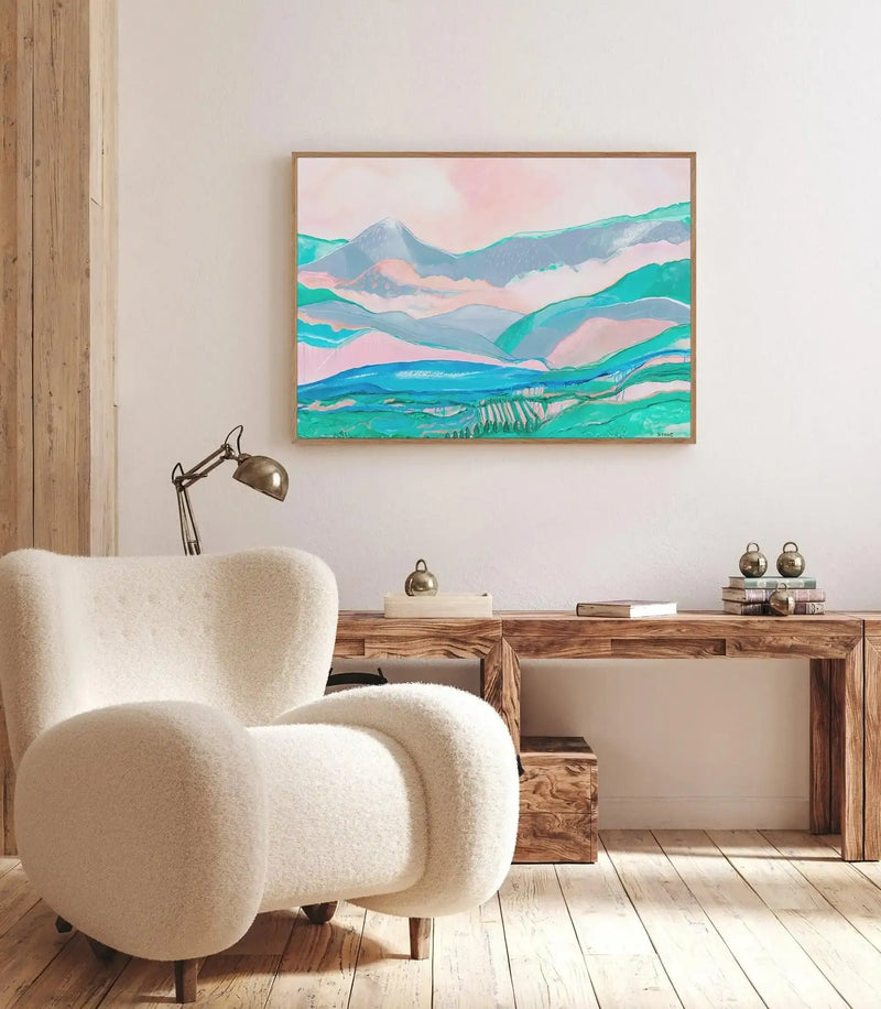 Lifestyle shot of a minimalist living room featuring a large framed canvas art print displayed above a wooden console table. The print is an abstract landscape with rolling hills and mountains in shades of teal, aqua, light blue, and soft pink, with a light wood frame. A cream-colored boucle armchair with wooden legs is positioned in the foreground to the left, and a vintage-style brass desk lamp sits on the console table. The room has light wooden floorboards and a white wall.