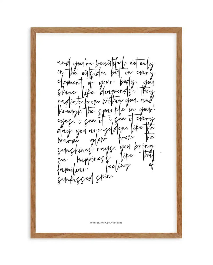 Framed art print featuring handwritten calligraphy on white background. The script reads a romantic poem beginning with "and you're beautiful, not only on the outside..." The flowing, cursive text in black ink fills most of the frame, with some words crossed out for artistic effect. At the bottom in small printed text: "YOU'RE BEAUTIFUL, I LOVE ET ORIEL". The artwork is set in a natural wood frame with a warm honey tone and simple rectangular profile.