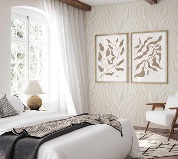 Bedroom styled with Zebra on Linen in Bone wallpaper covering the walls, featuring a subtle cream and beige wavy stripe pattern with a woven texture. Two framed botanical prints with taupe leaf designs hang on the wallpapered wall in gold frames. A white bed with gray and cream bedding sits in the foreground, a wooden mid-century chair in the background right, windows with white curtains on the left, and a round textured lamp on the windowsill.