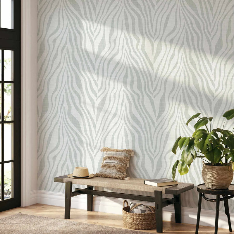 Zebra on Linen in Mist wallpaper displayed on an interior wall, showing a repeating geometric pattern of vertical wavy stripes in soft gray and cream tones creating an interlocking leaf or flame-like motif. The wallpaper is shown installed in a bright, minimalist entryway with natural light streaming through a black-framed glass door, styled with a dark wooden bench, woven basket, straw hat, decorative pillow, potted plant in a textured planter, and a bentwood chair, all on light wood flooring.