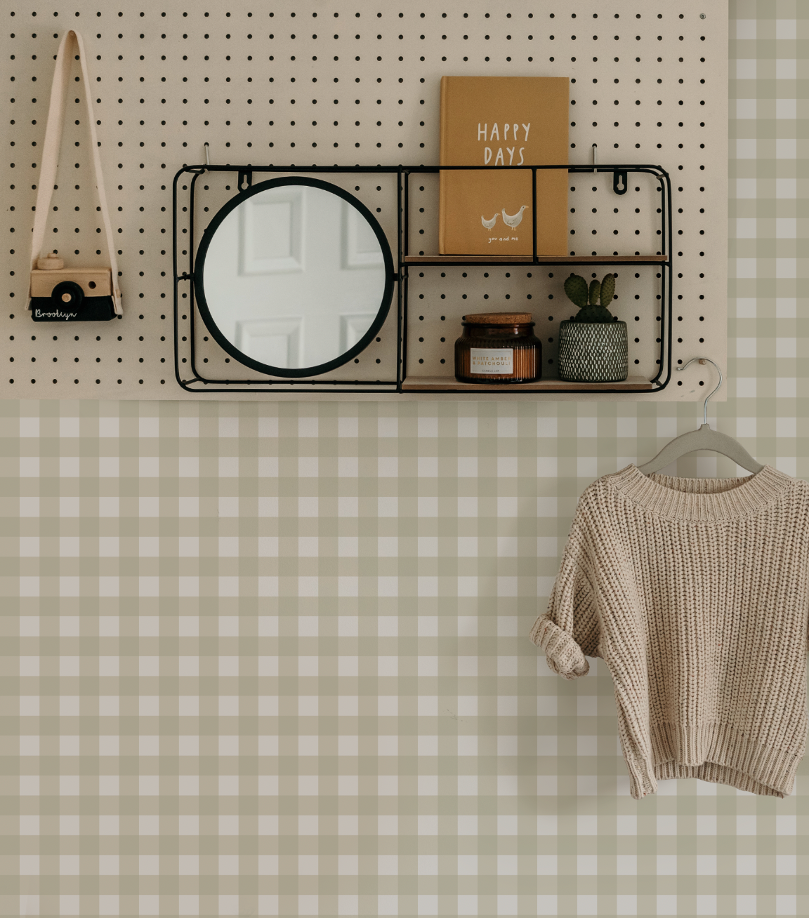 Lifestyle shot featuring a small gingham check wallpaper in a new neutral green colour. The wallpaper covers the lower half of a wall, with a cream-coloured pegboard above it. On the pegboard, a black wire shelf with a round mirror and two wooden shelves displays a book titled "HAPPY DAYS," a candle, and a small potted cactus. A small wooden toy camera hangs from the pegboard. A light beige knitted sweater hangs on a grey hanger against the gingham wallpaper.
